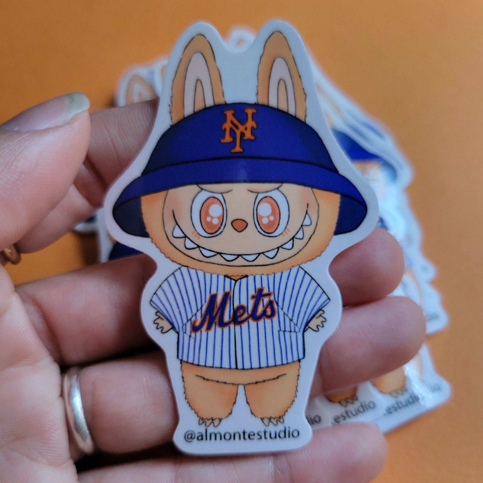 Hand holding a cartoon character sticker with a blue cap and 'Mets' jersey on an orange background