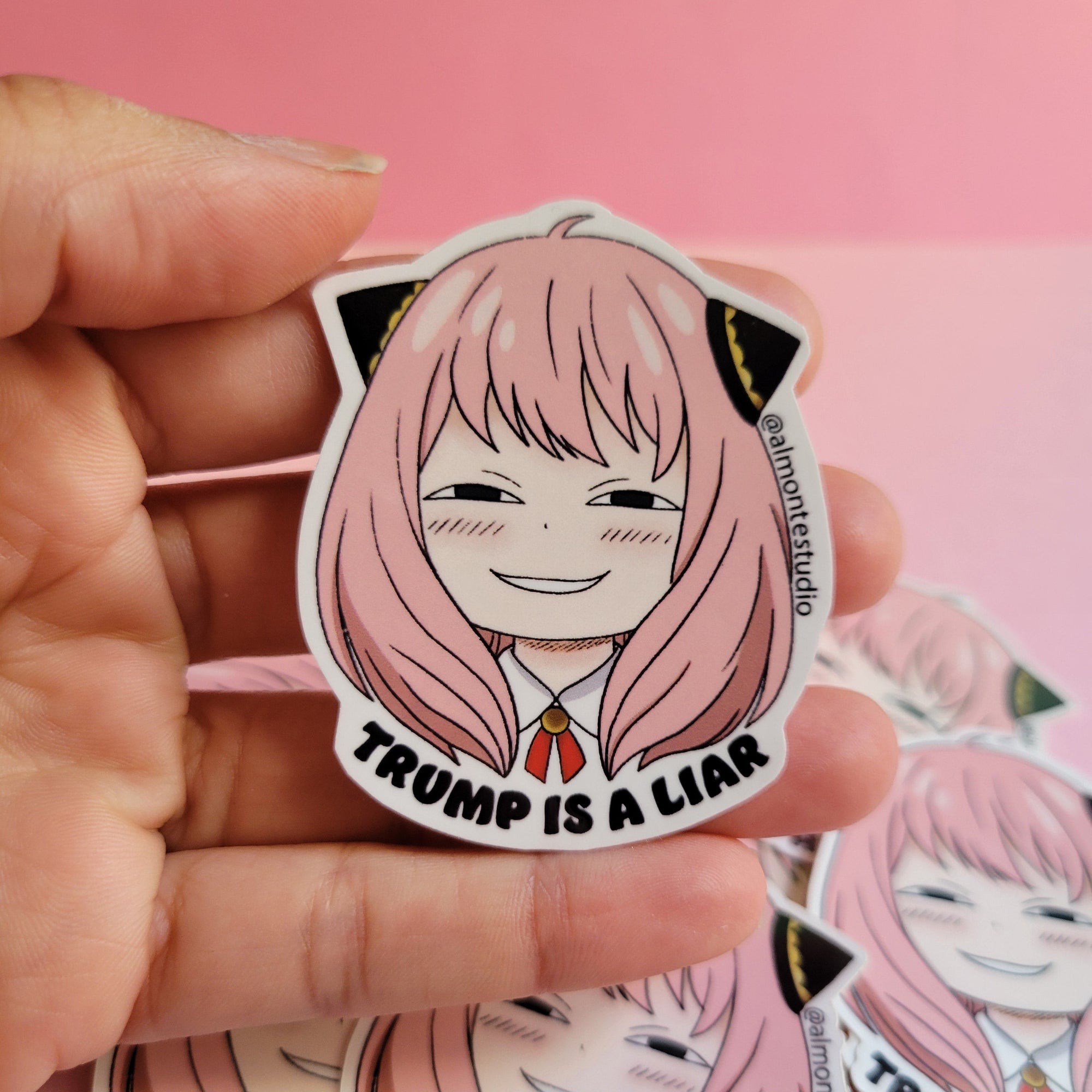 Sticker with an anime character and text 'TRUMP IS A LIAR' held by a hand against a pink background.