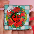 Hand holding a colorful strawberry-themed pin with a checkered border against a pink background