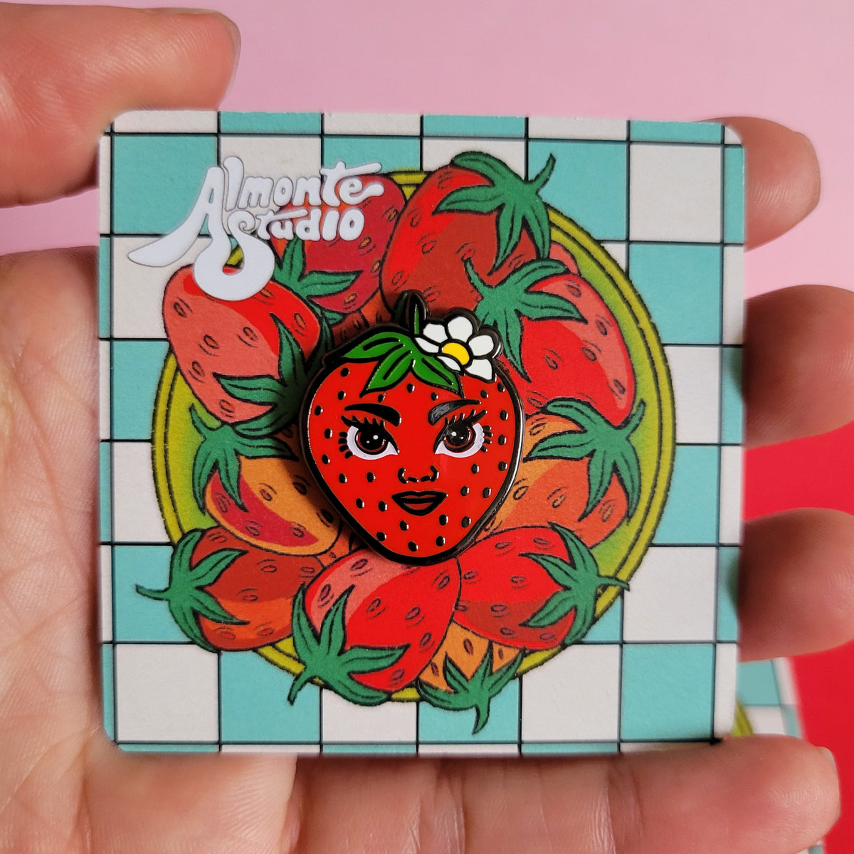 Hand holding a colorful strawberry-themed pin with a checkered border against a pink background