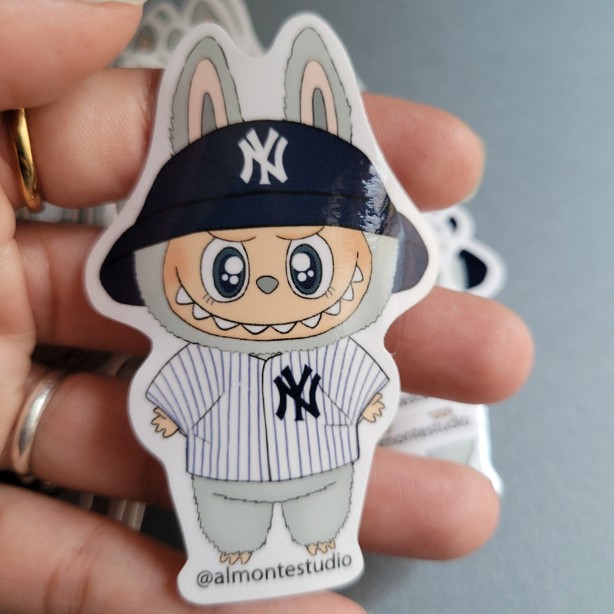 Hand holding a sticker of a cartoon character wearing a New York Yankees cap against a gray background