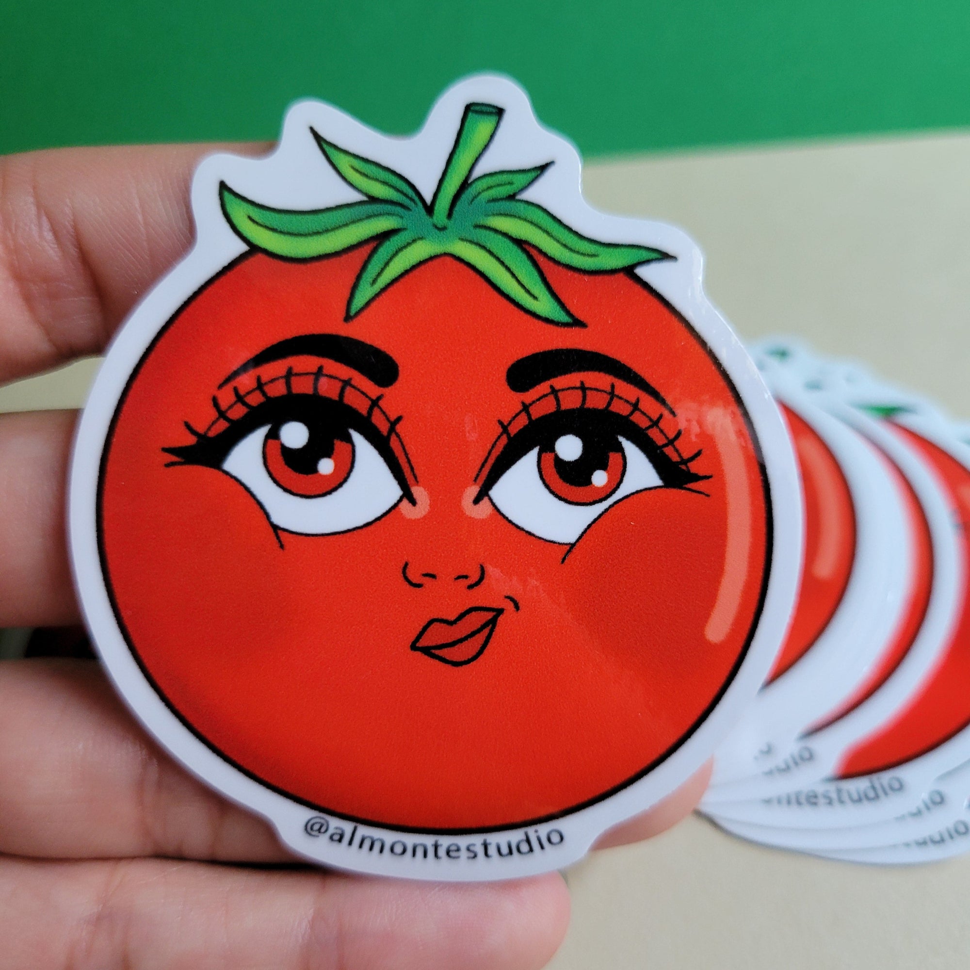 Hand holding a sticker with a stylized face design on a green and beige background