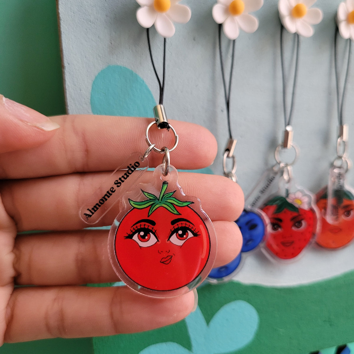 Hand holding a keychain with a tomato design, with a colorful background of similar keychains.