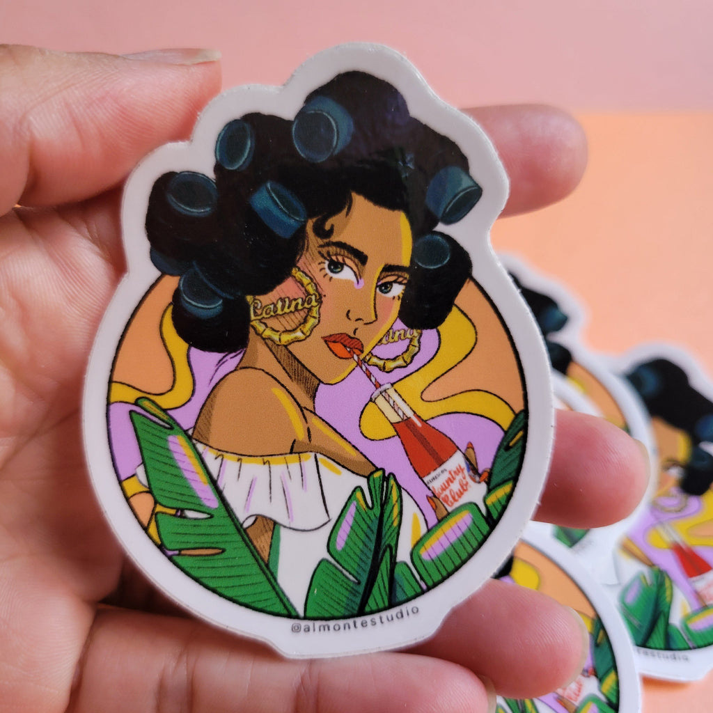Hand holding a colorful sticker with a stylized illustration of a woman with black hair, a yellow top, and green leaves on a pink and orange gradient background.