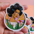 Hand holding a colorful sticker with a stylized illustration of a woman with black hair, a yellow top, and green leaves on a pink and orange gradient background.