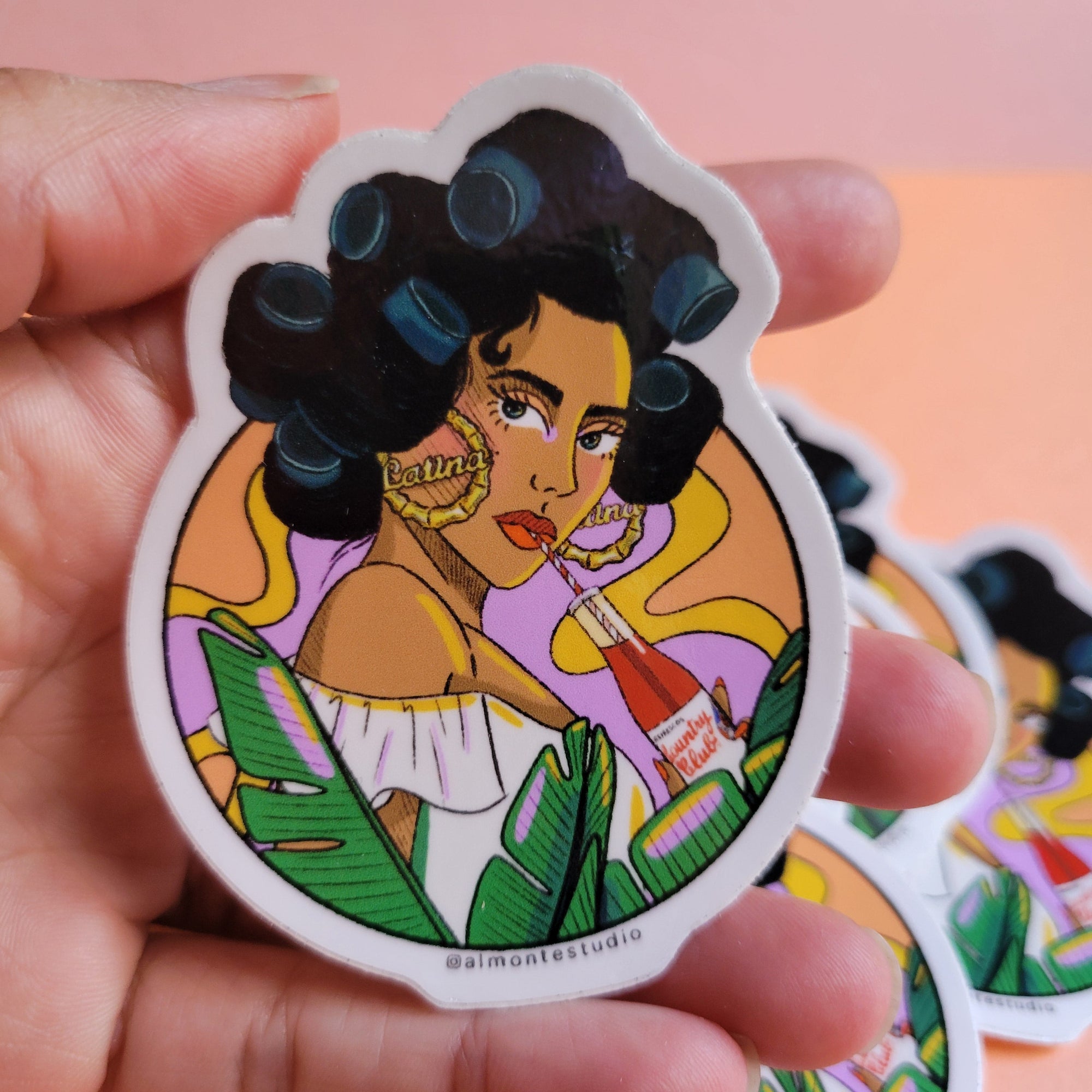 Hand holding a colorful sticker with a stylized illustration of a woman with black hair, a yellow top, and green leaves on a pink and orange gradient background.