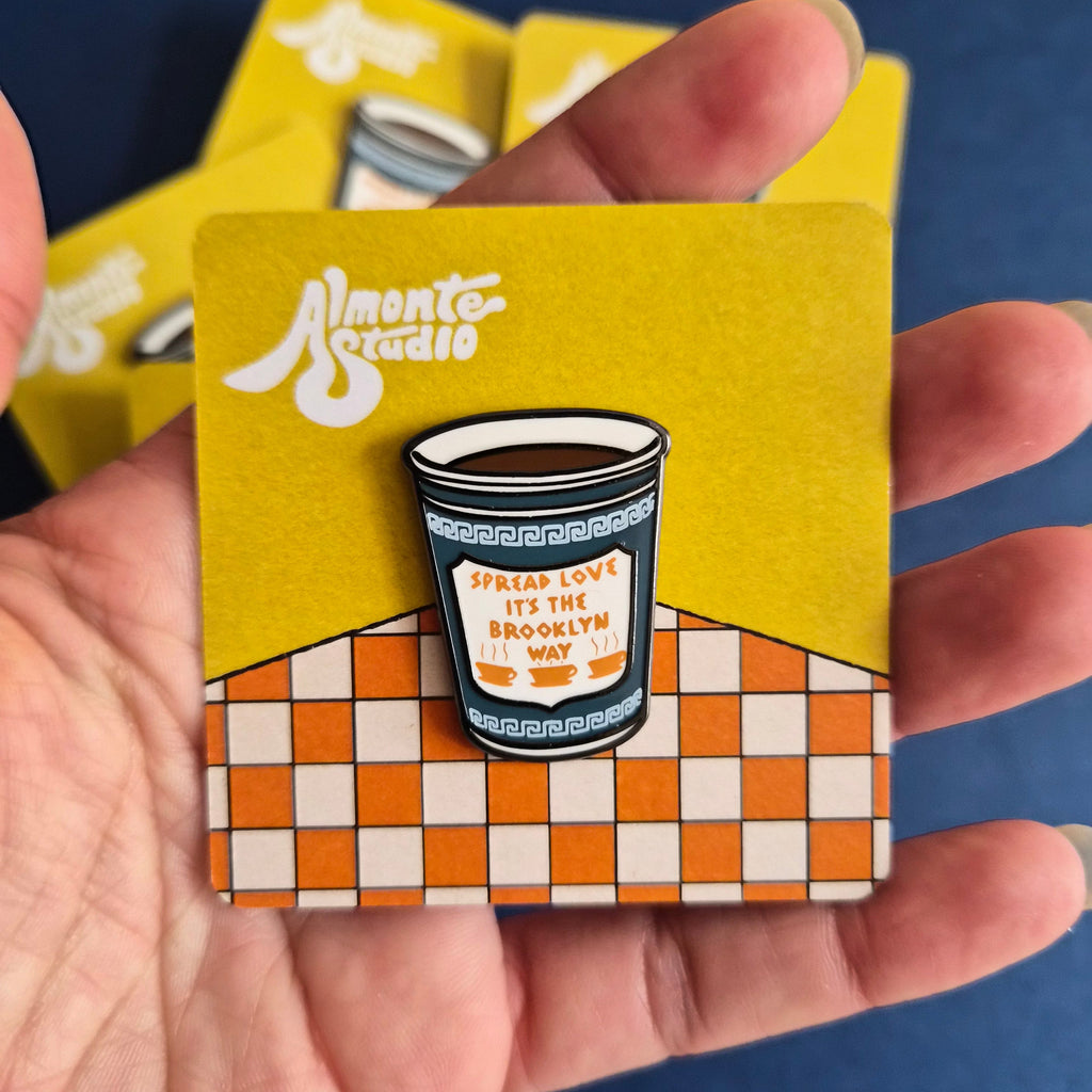 A hand holding a small enamel pin shaped like a coffee cup, mounted on a yellow and orange checkered backing card. The card has the Almonte Studio logo printed on it, and other cards are visible in the background against a dark blue surface.