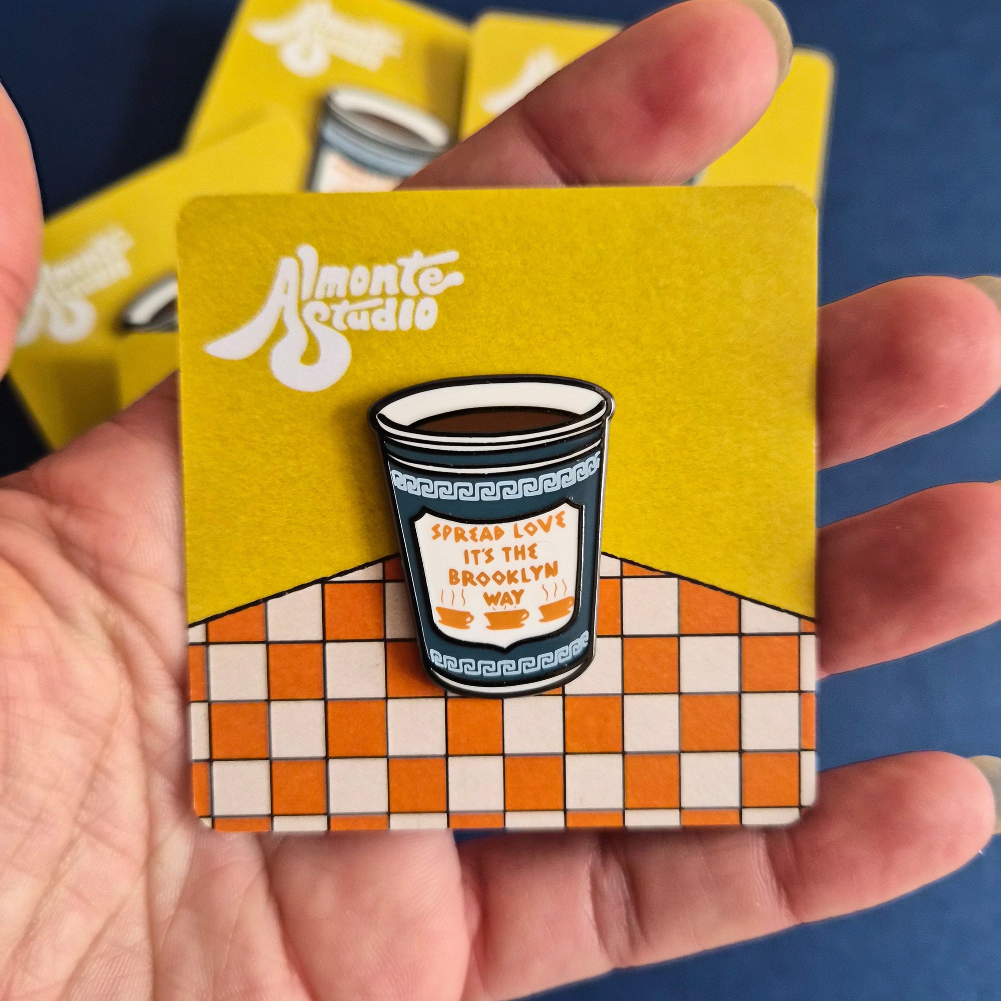 A hand holding a small enamel pin shaped like a coffee cup, mounted on a yellow and orange checkered backing card. The card has the Almonte Studio logo printed on it, and other cards are visible in the background against a dark blue surface.