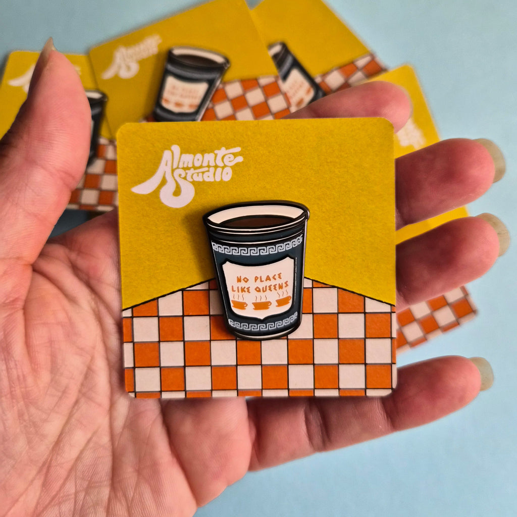 A hand holds several small square cards, each featuring a coffee cup enamel pin mounted on a yellow backing card with a checkered pattern. The main card in front shows the brand name 'Almonte Studio in white script.